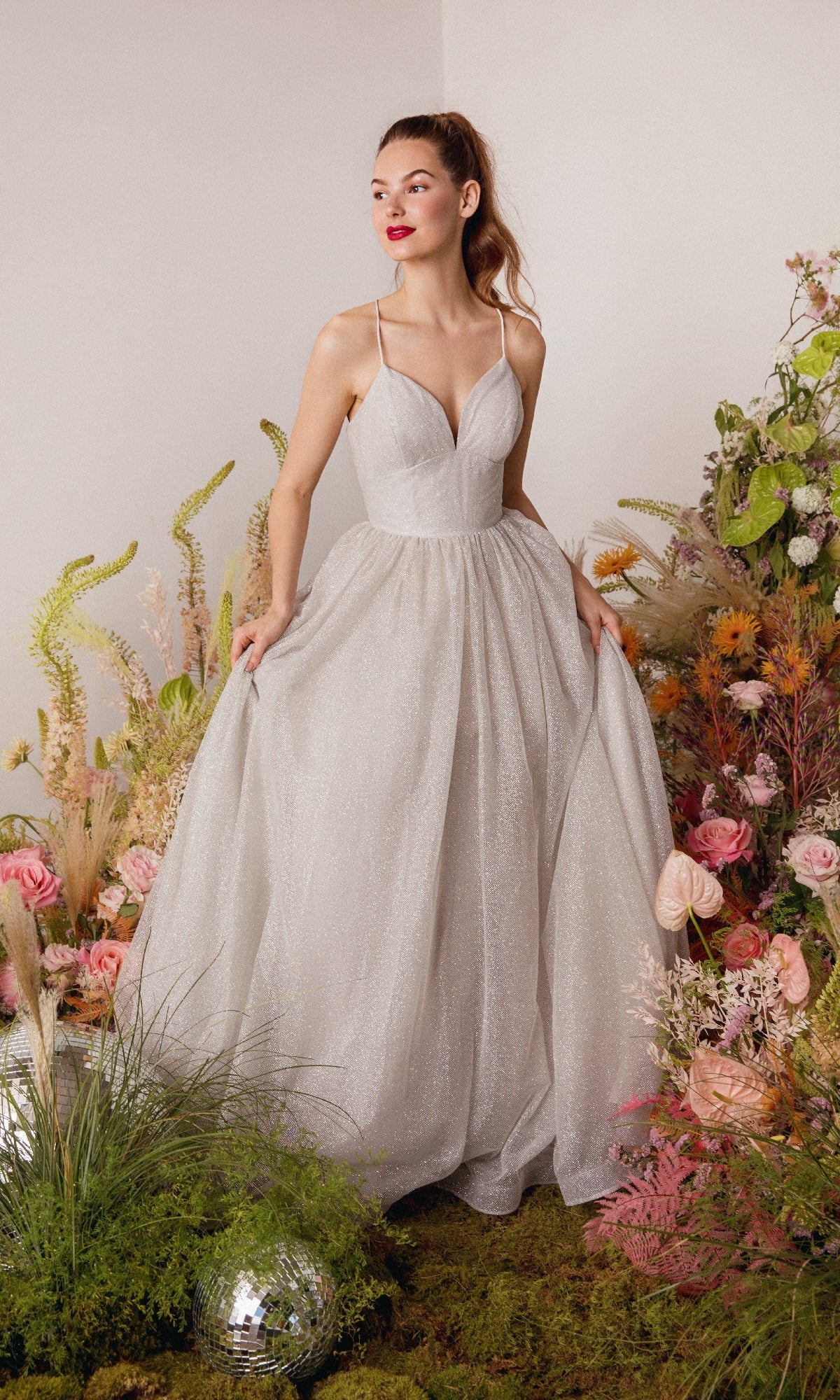 A woman in the Strappy-Back Glitter Prom Ball Gown: Eureka 9009 stands among colorful flowers and greenery, holding the sides of her sparkling gown.